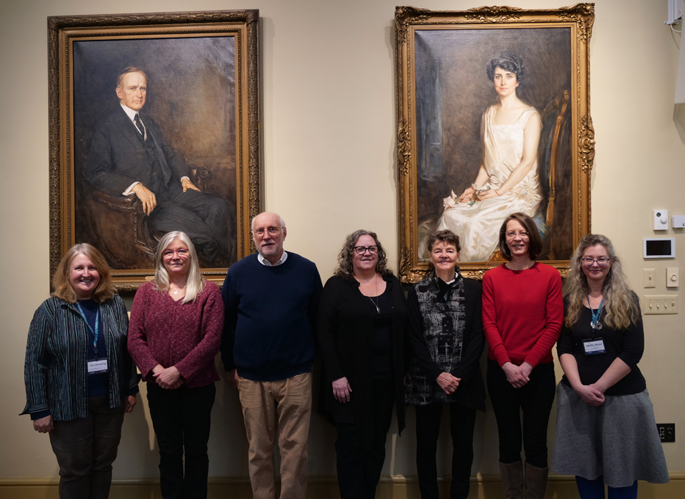 Photo of 7 adults standing side by side in front of portraits of Calvin and Grace Coolidge. Left to right: Lisa Downing, Anne Teschner, Russell Carrier, Katy Wight, Debin Bruce, Ruth Francis, Molly Moss