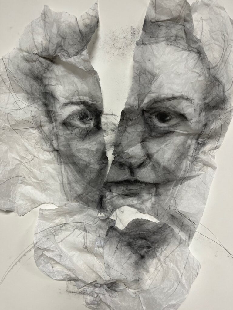 A charcoal drawing of face done on translucent paper which is ripped down the center of the face and across the chin, revealing a smudged white surface below. 