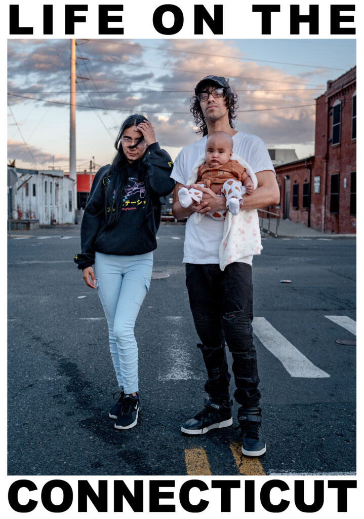 A photograph of a young woman and a young man, standing together with a streetscape behind them. The man holds an infant. Dramatic clouds hang in the sky over aging buildings. 