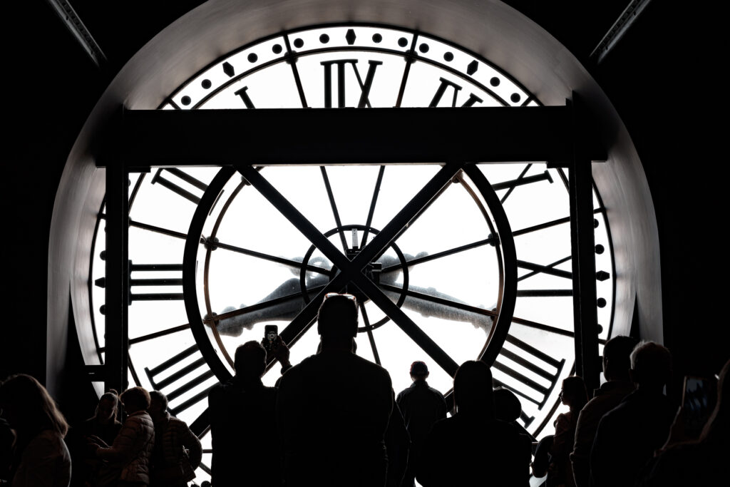 Photography depicts a crowd of people looking out from within a clock tower whose clock face bathes the people in light. 