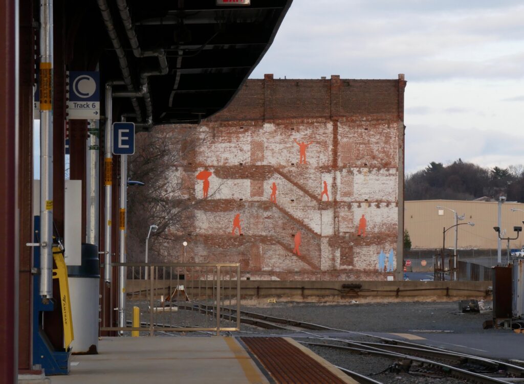 A train station with a brick building in the background, decorated with a mural showing human silhouettes in orange and blue. 