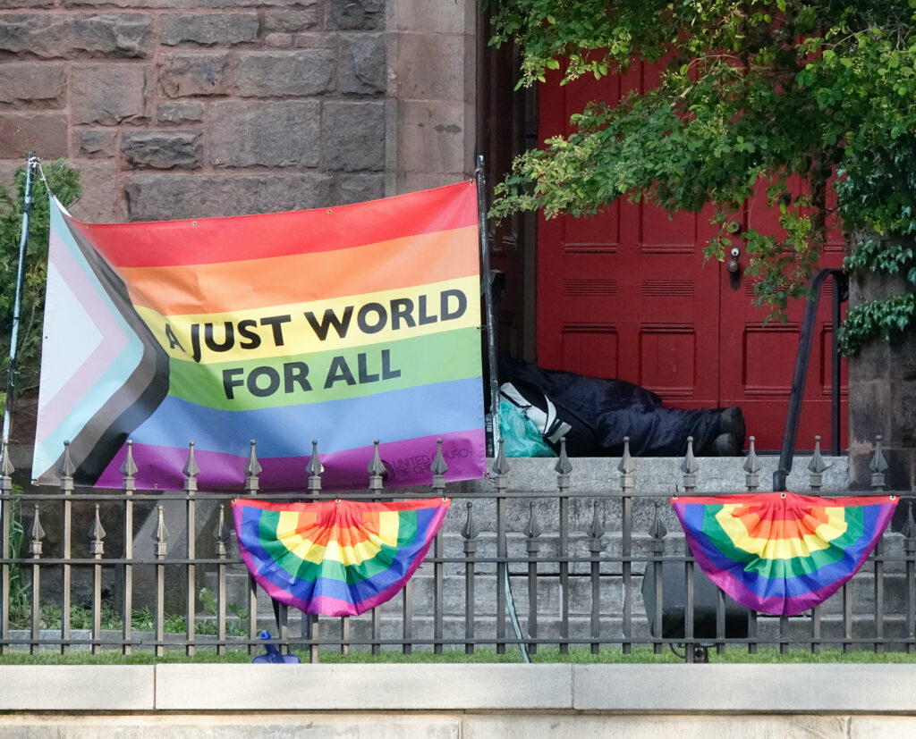 Color photograph of the First Churches building's red door, with a Queer Pride Flag adorning the front gate. The flag reads, "A just world for all." A sleeping person reclines in the door way in a black sleeping bag.