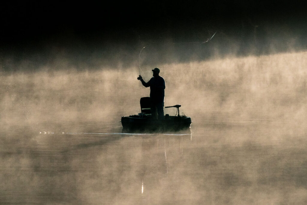 A person in a small boat casts a fly across the river as fog rises. 