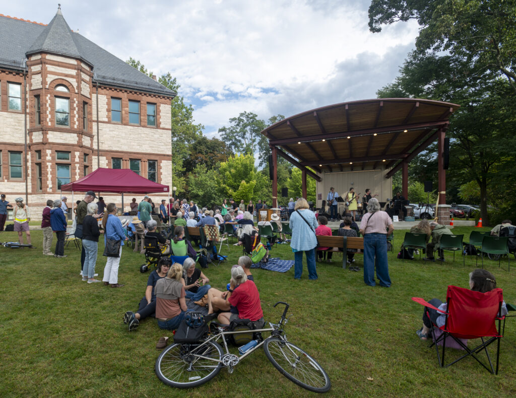 An outdoor concert with four musicians on a large stage. In the audience, people sit on the ground and in chairs, watching the band and chatting amongst themselves. 