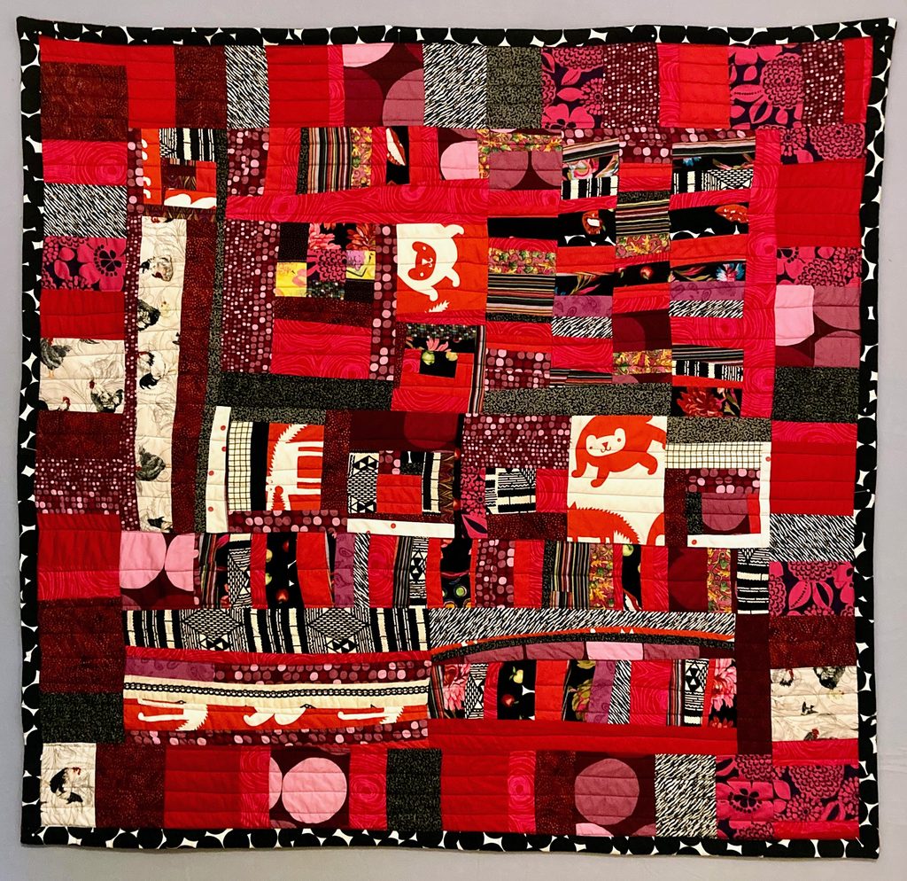A square quilt of improvised square and rectangular patchwork made of various red tones with accents of white, cream, and black. 